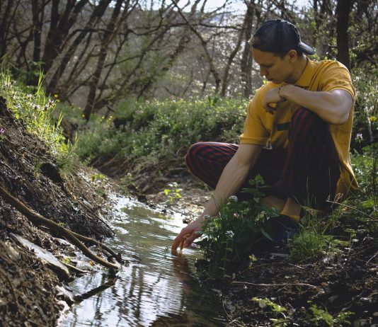A young man is kneeling in front of a small stream in the woods.
